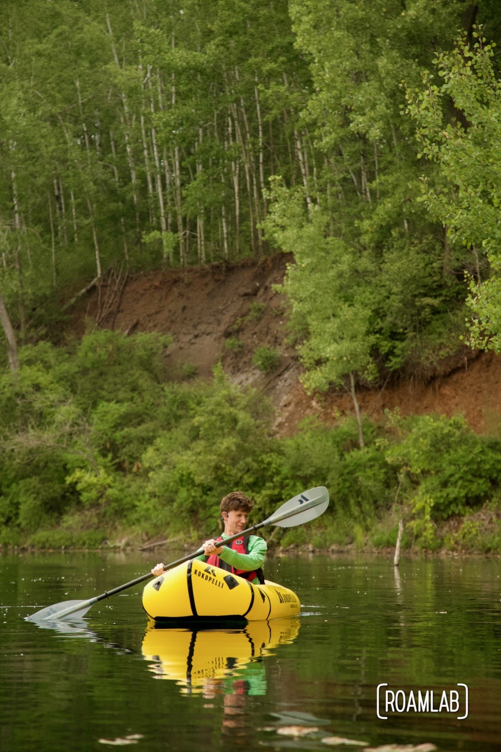 Man in a yellow raft paddling along forested red earth cliffs in Cuyuna Country State Recreation Area.