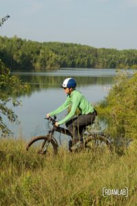 Man on a black mountain bike, cycling through tall grass by a forested lake.