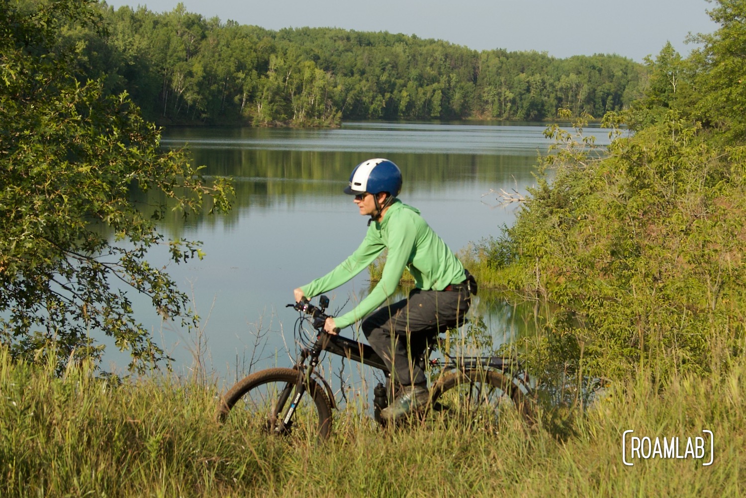 Man on a black mountain bike, cycling through tall grass by a forested lake.
