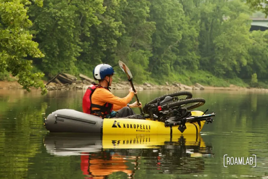 Man in a yellow raft with a bicycle strapped to the front paddling down a river.