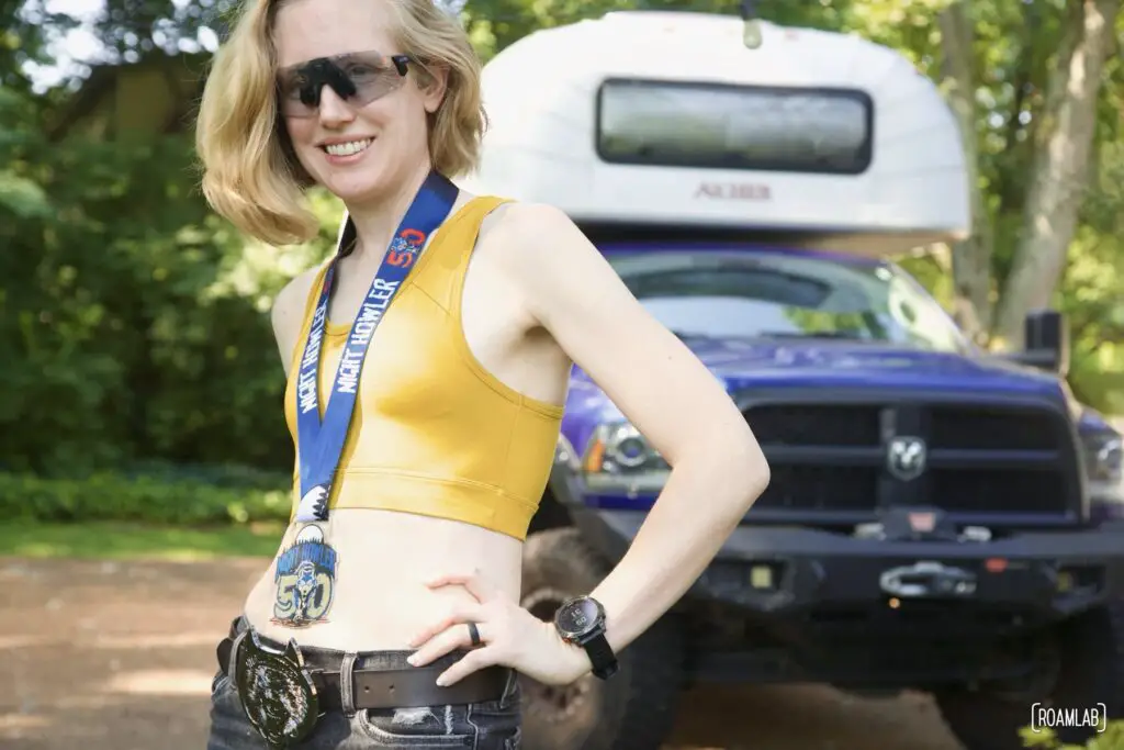 Woman standing in front of a truck camper with wolf head belt buckles and a medal for the Night Howler 50.