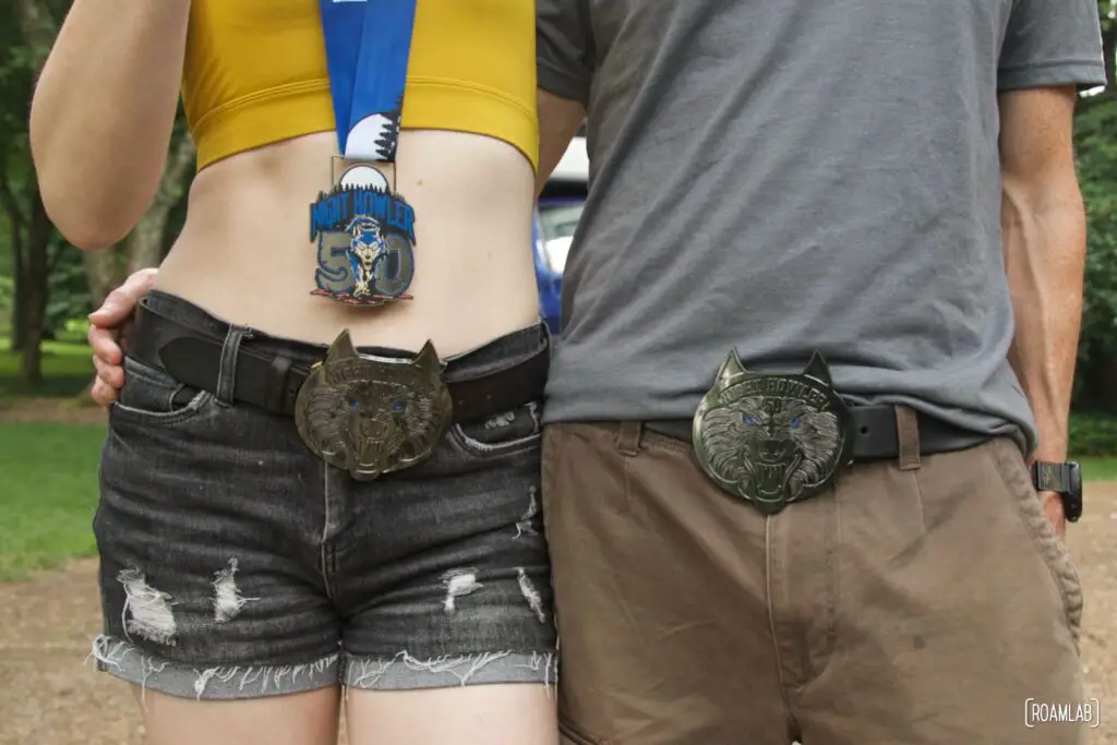Closeup of the waistline of a man and woman standing together with wolf head belt buckles and a medal for the Night Howler 50.